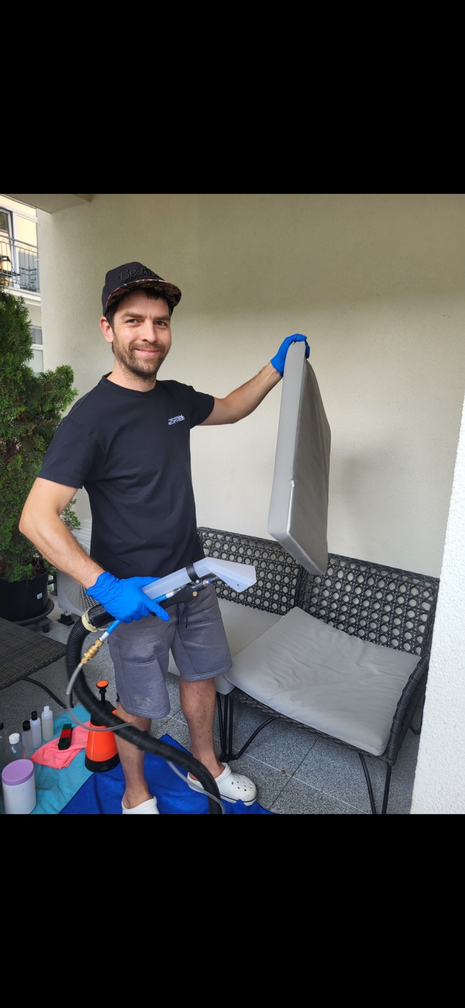 Mężczyzna w czarnej koszulce i niebieskich rękawiczkach, trzymający wyprany, szary materac od sofy na tle balkonu z rattanowymi meblami i roślinami, obok leży sprzęt do czyszczenia.