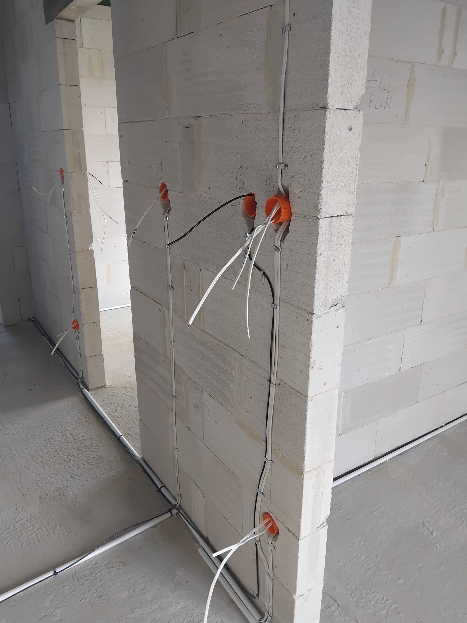 Surowy stan instalacji elektrycznej w nowym budynku: widoczne przewody prowadzone po ścianach z betonu komórkowego, pomarańczowe puszki instalacyjne, kable w peszlach na podłodze, napis 'ALI TOK'...