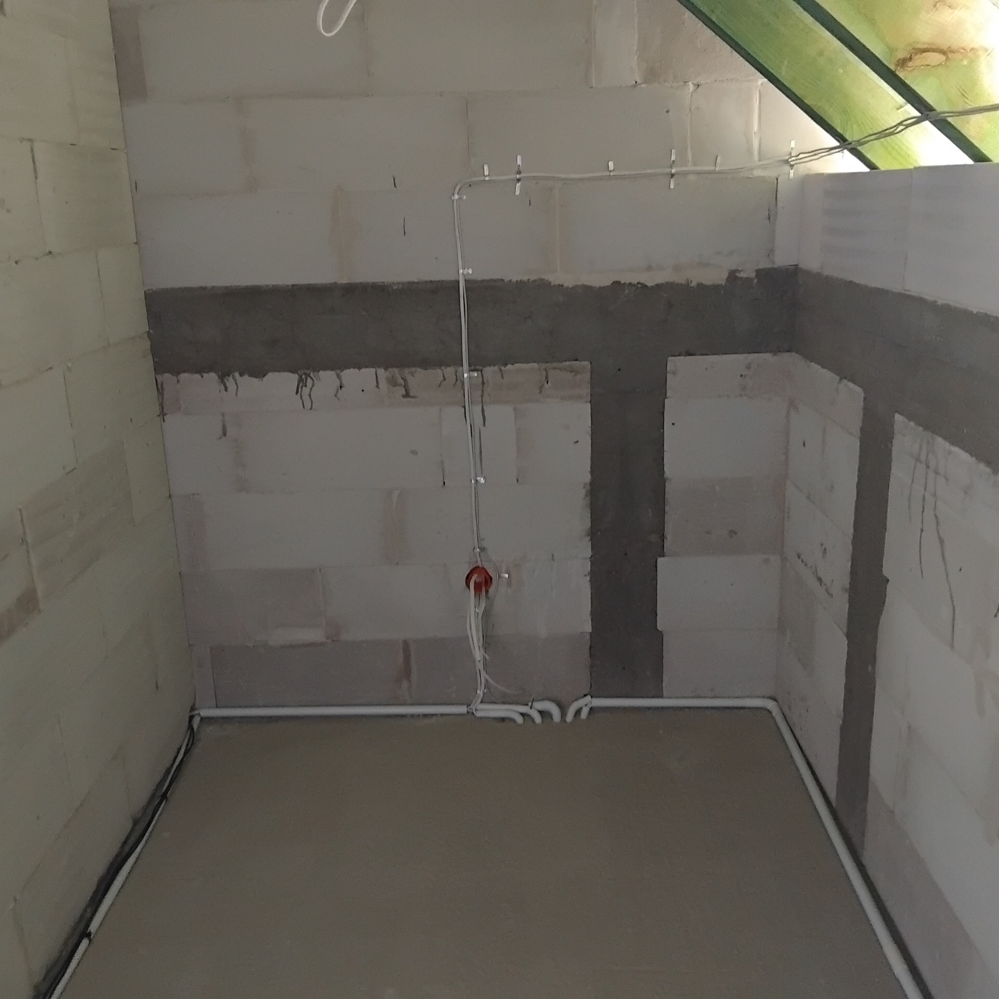 Surowy stan pomieszczenia w trakcie prac: widoczne białe ściany z betonu komórkowego z częściowo nałożoną szarą zaprawą, świeża wylewka na podłodze i poprowadzone kable elektryczne w białych...