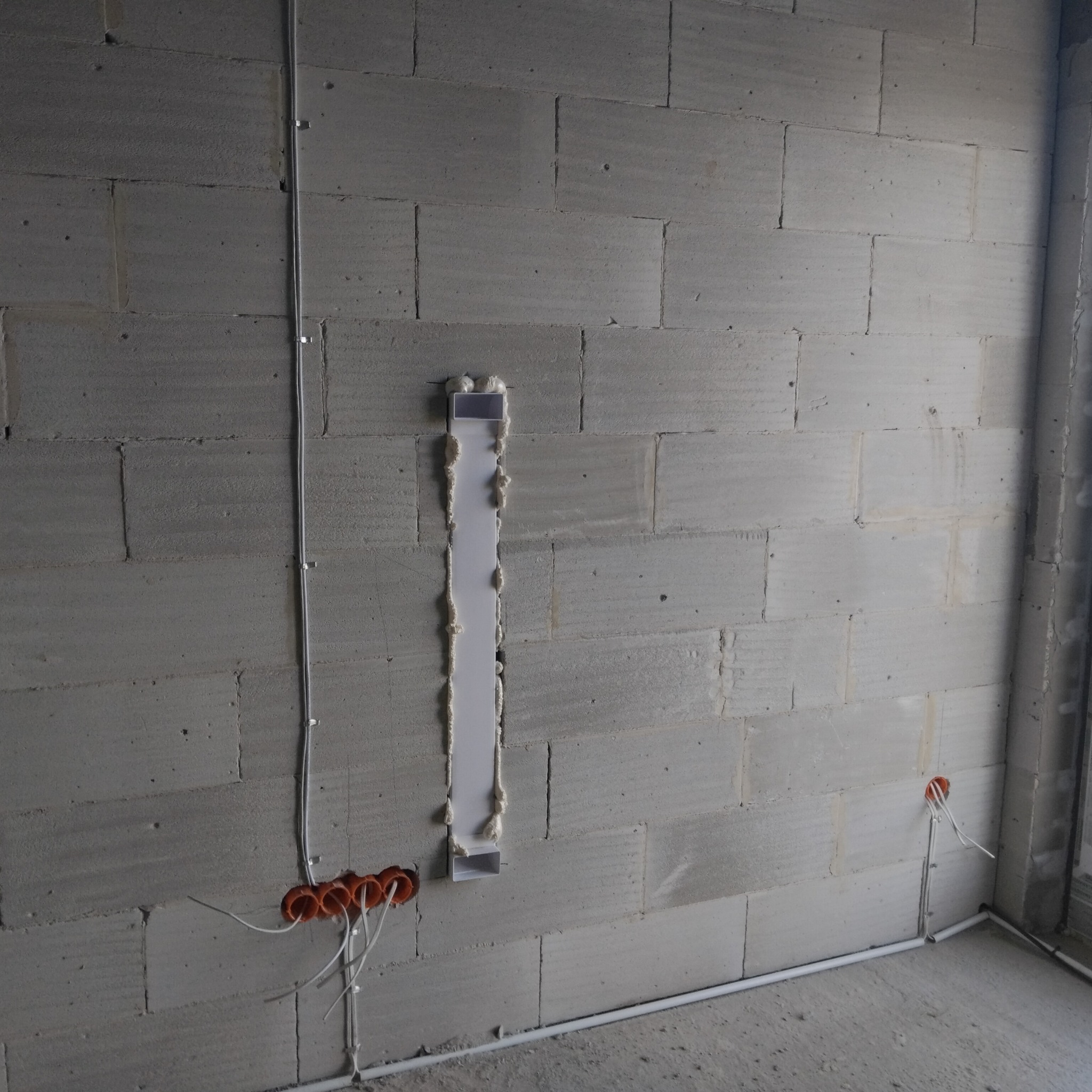 Szara ściana z betonu komórkowego w trakcie prac instalacyjnych, widoczne pomarańczowe puszki elektryczne, białe przewody oraz pionowy kanał kablowy z pianką montażową.