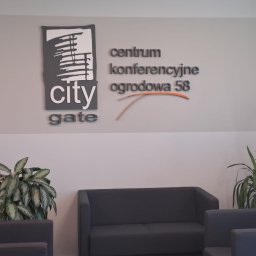 Akademia Marketingu KFK - Wnętrze centrum konferencyjnego z logo firmy City Gate na ścianie, szarymi sofami i roślinami doniczkowymi.
