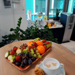 Akademia Marketingu KFK - Stół konferencyjny z paterą owoców: truskawki, winogrona, pomarańcze, banany, filiżanka cappuccino i ciastko na tle okna biurowego.