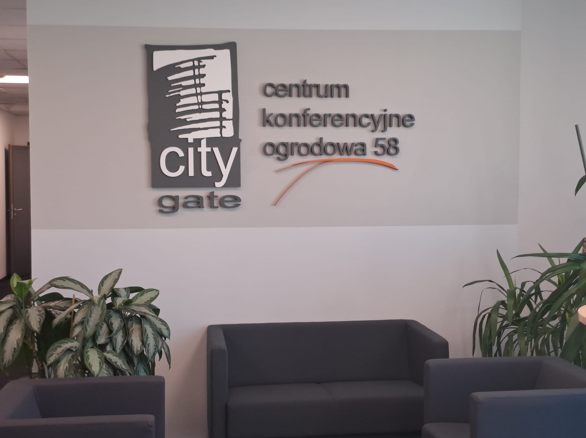 Wnętrze centrum konferencyjnego z logo firmy City Gate na ścianie, szarymi sofami i roślinami doniczkowymi.