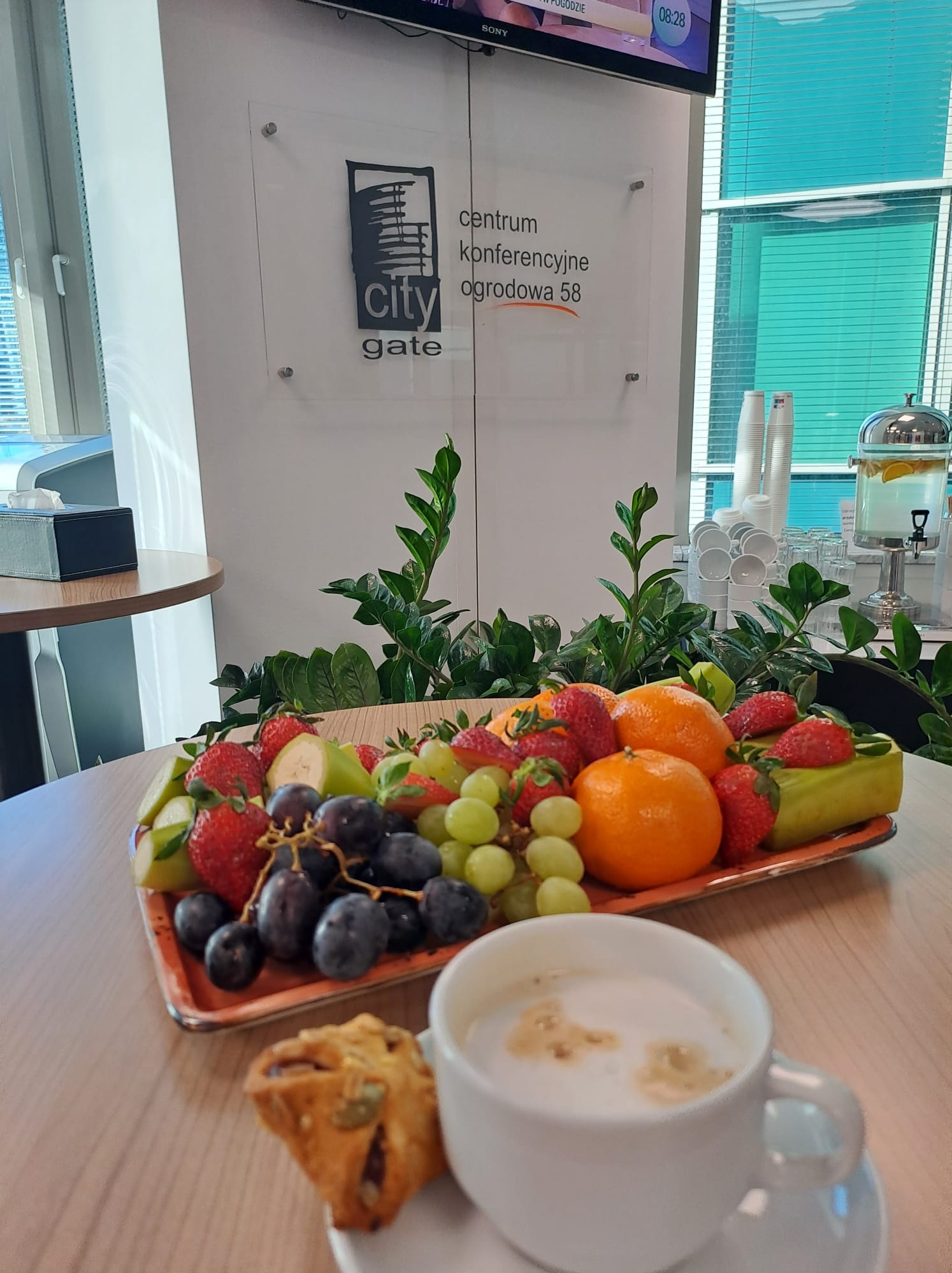 Stół w sali konferencyjnej z owocami, ciastkiem i kawą, w tle logo centrum konferencyjnego City Gate i dystrybutor wody z cytryną.