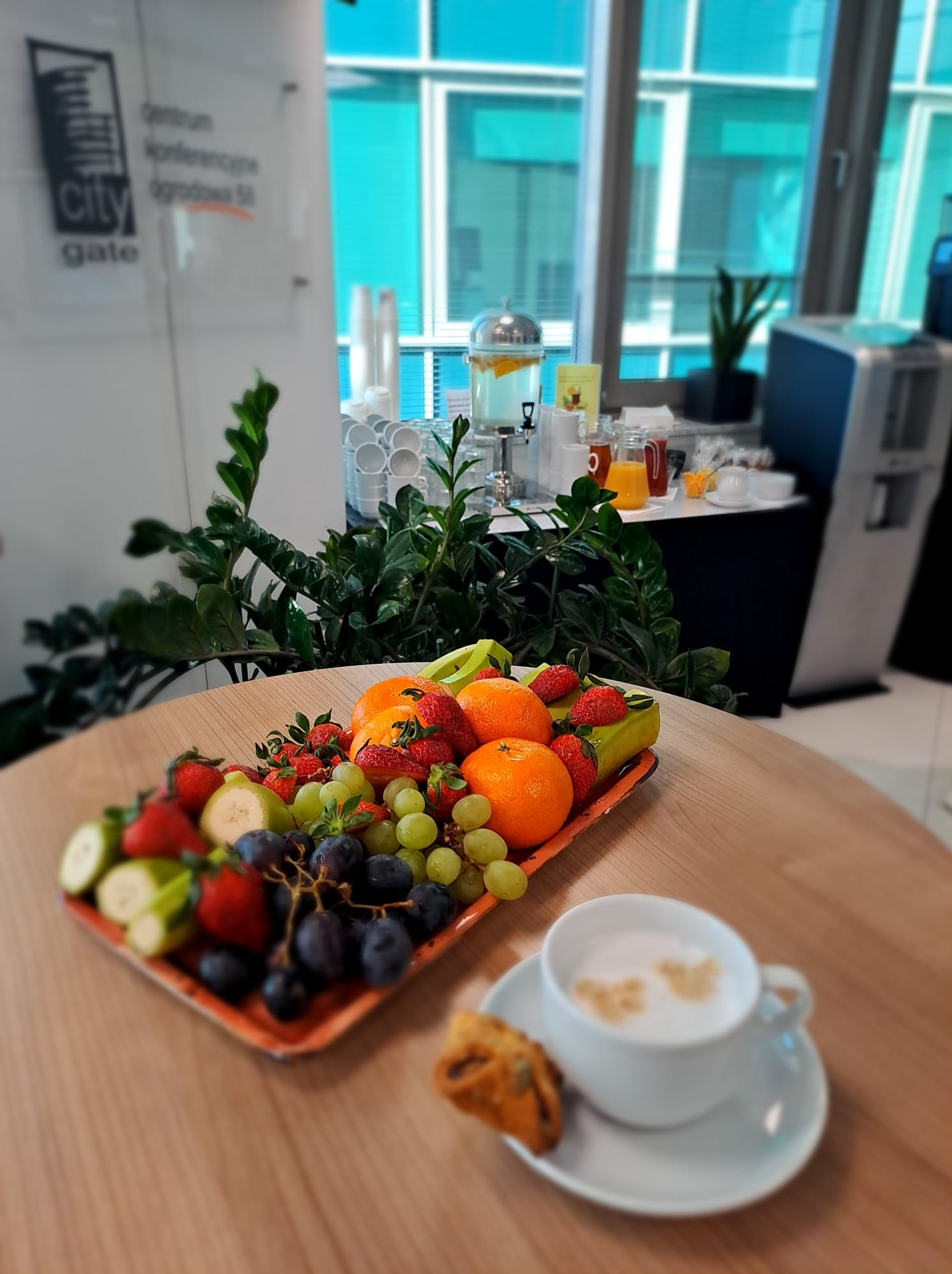 Stół konferencyjny z paterą owoców: truskawki, winogrona, pomarańcze, banany, filiżanka cappuccino i ciastko na tle okna biurowego.