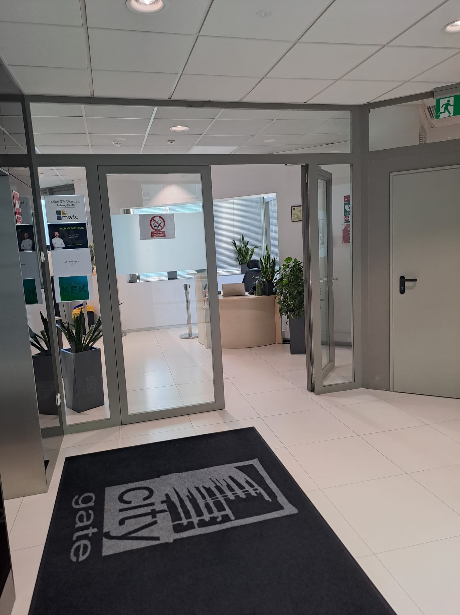 Wejście do biura z widocznym logo 'gate City' na wycieraczce, przez szklane drzwi widać recepcję i plakaty informacyjne.