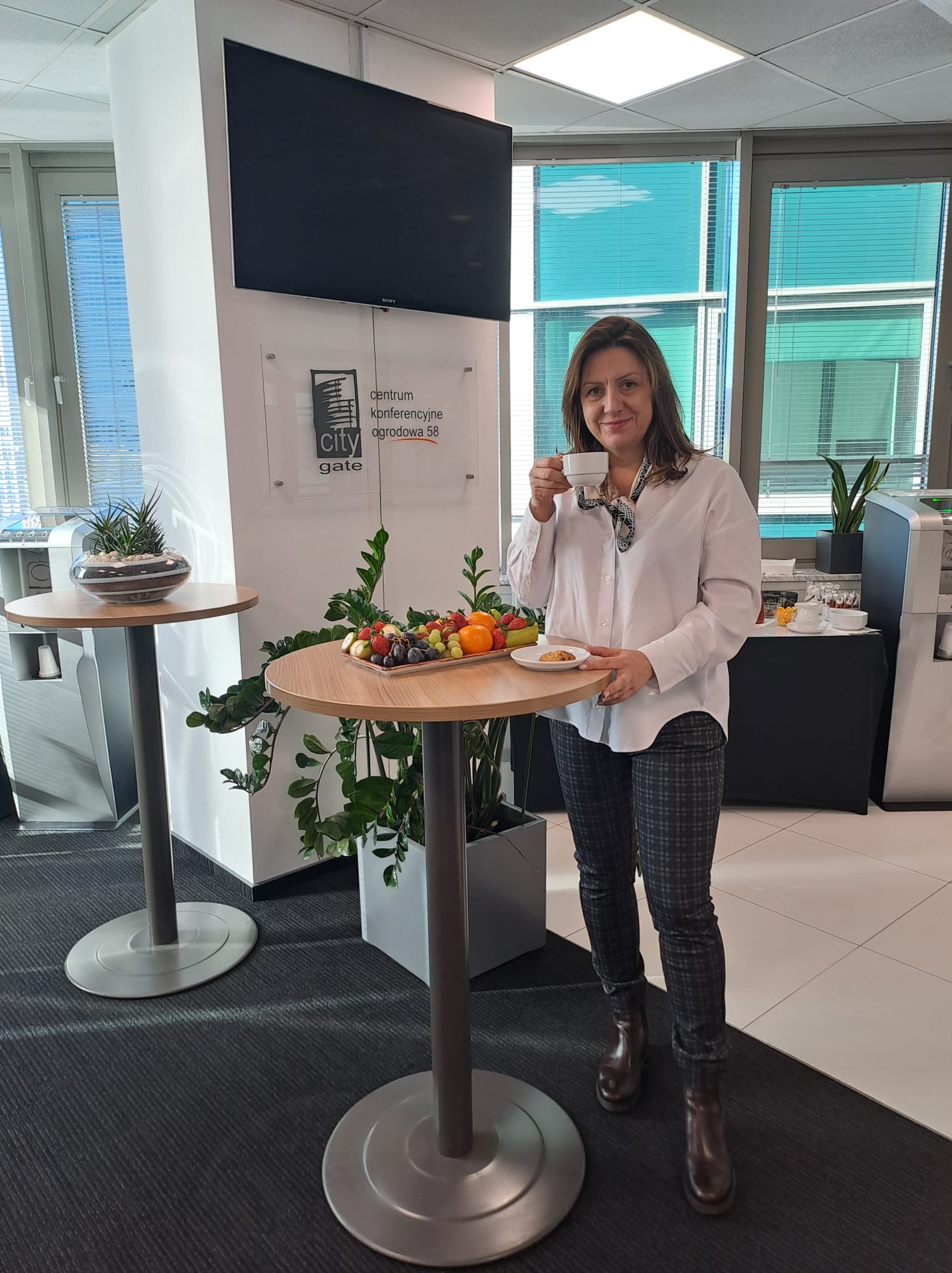 Kobieta w białej koszuli pije kawę podczas przerwy na szkoleniu w centrum konferencyjnym City Gate, z przekąskami na stole. Widoczne okna i monitor.
