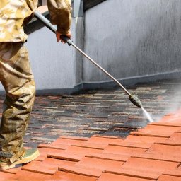 BS USŁUGI WYSOKOŚCIOWE - Częściowy widok osoby w kombinezonie czyszczącej dach z czerwonej dachówki za pomocą myjki ciśnieniowej. Widoczny efekt czyszczenia - różnica koloru dachówek.