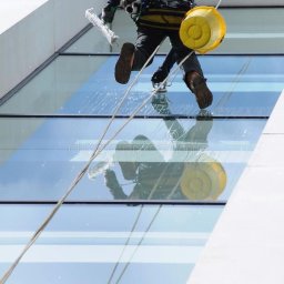 BS USŁUGI WYSOKOŚCIOWE - Pracownik na linach myje okna wieżowca za pomocą ściągaczki. Widoczne odbicie w oknach i żółte wiadro. Praca na wysokościach.