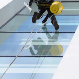 Pracownik na linach myje okna wieżowca za pomocą ściągaczki. Widoczne odbicie w oknach i żółte wiadro. Praca na wysokościach.