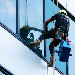 BS USŁUGI WYSOKOŚCIOWE - Pracownik w uprzęży, myje okno na wysokości. Widoczne narzędzia pracy i fragment umytego okna. Budynek biurowy z refleksami nieba.
