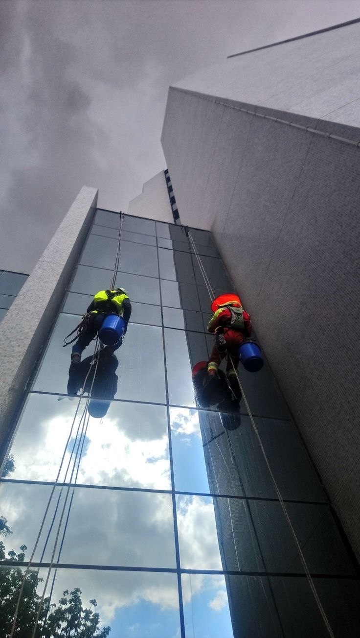 Dwóch alpinistów w kaskach i uprzężach, z wiadrami, myje szklaną fasadę wysokiego biurowca. W oknach odbija się pochmurne niebo.