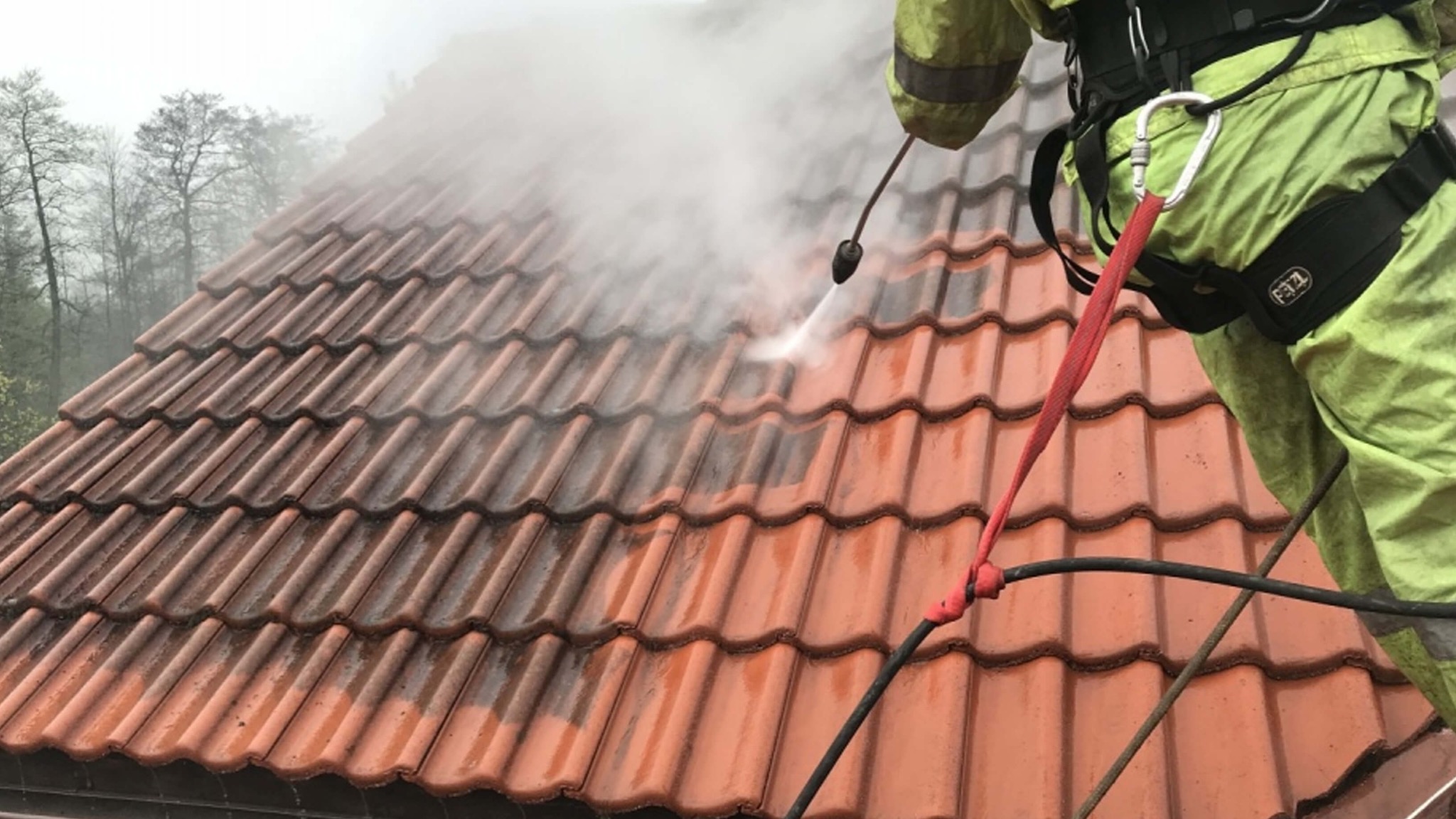 Częściowy widok dachu z czerwonej dachówki, czyszczonego pod ciśnieniem przez pracownika w uprzęży. Połowa dachu umyta, druga brudna. Widoczne drzewa w tle.