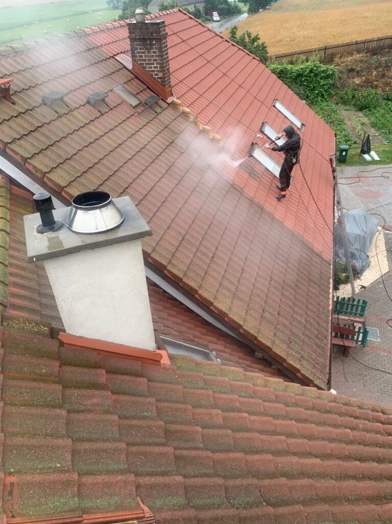 Częściowe mycie dachu z czerwonej dachówki, widoczne okna dachowe i komin. Pracownik w uprzęży czyści dach myjką ciśnieniową, wyraźny kontrast między czystą a zabrudzoną powierzchnią.