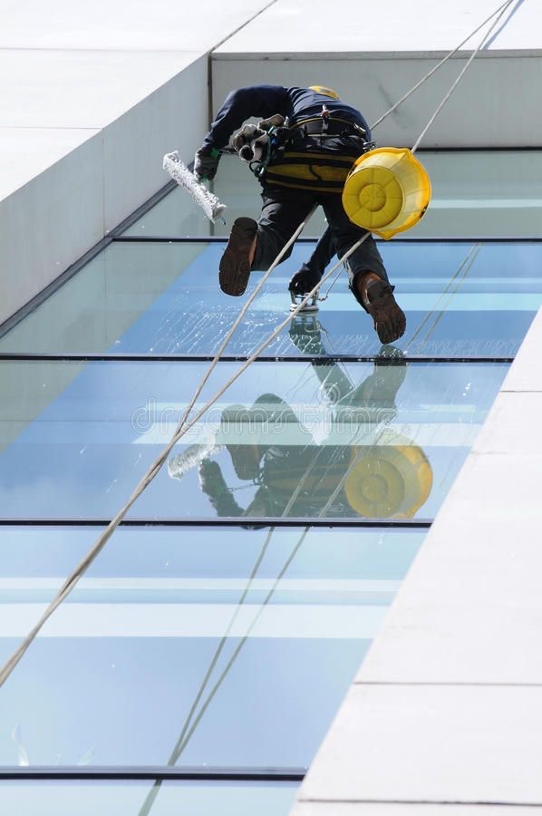Pracownik na linach myje okna wieżowca za pomocą ściągaczki. Widoczne odbicie w oknach i żółte wiadro. Praca na wysokościach.