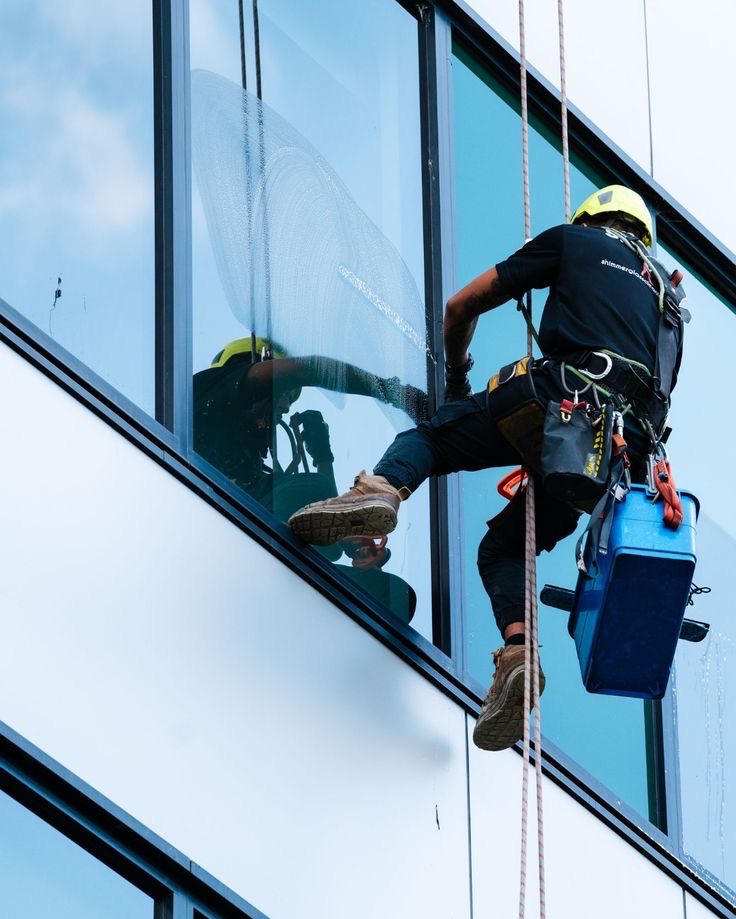 Pracownik w uprzęży, myje okno na wysokości. Widoczne narzędzia pracy i fragment umytego okna. Budynek biurowy z refleksami nieba.