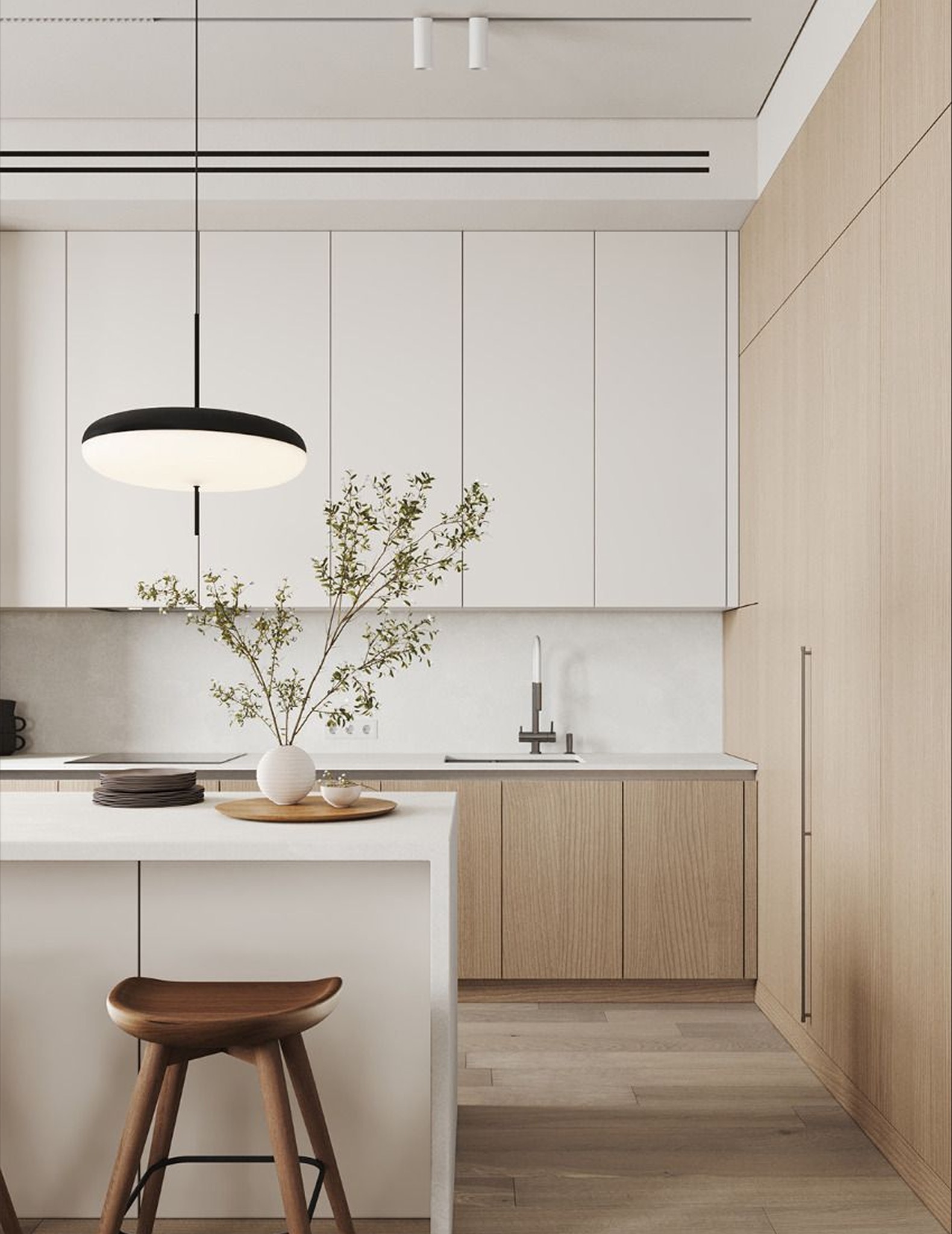 Minimalistyczna kuchnia z białymi szafkami górnymi i drewnianymi dolnymi, wyspa z hokerem, okrągła lampa wisząca. Wazon z gałązkami na blacie.