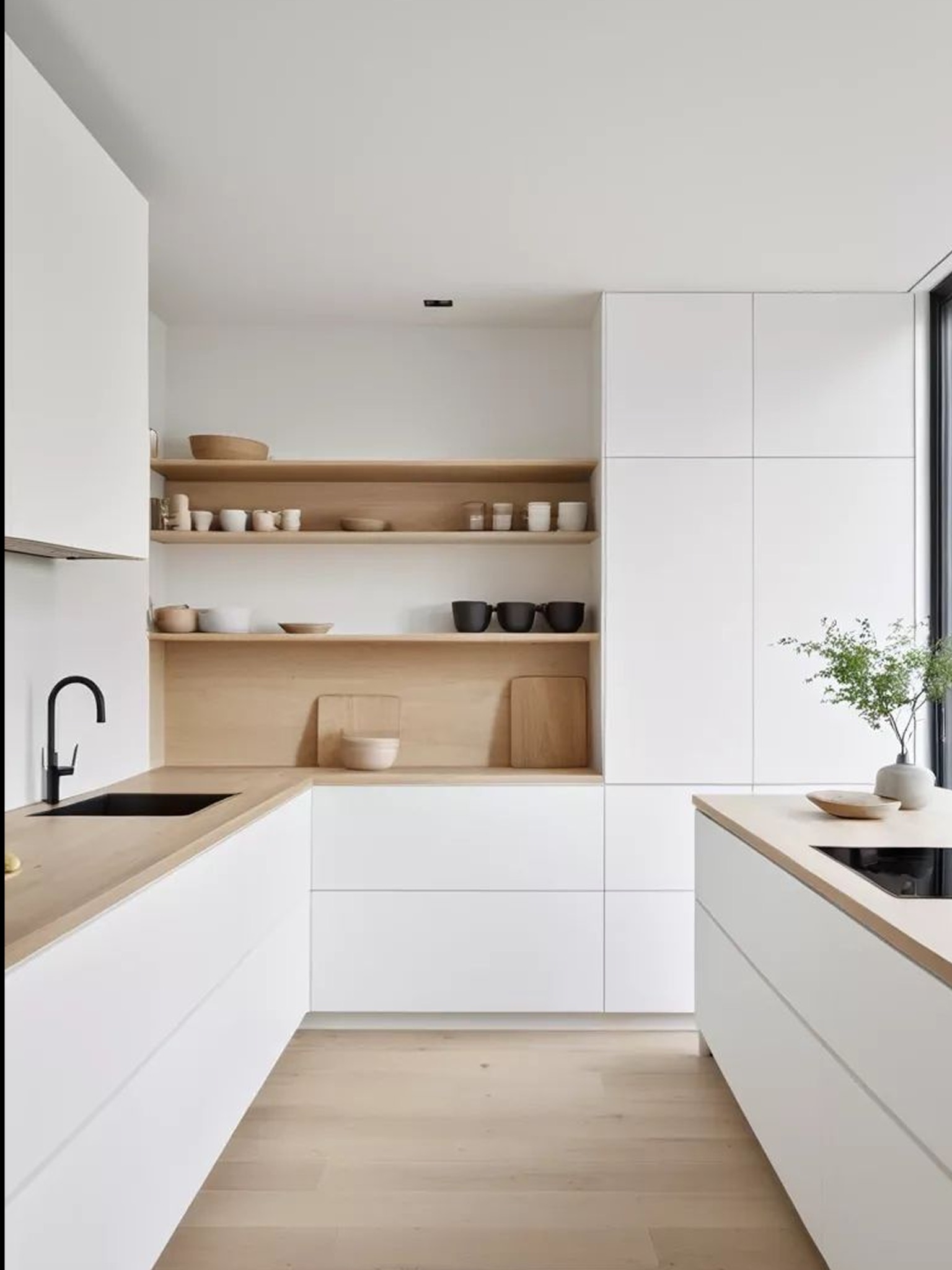 Minimalistyczna kuchnia: białe szafki, drewniany blat i półki z naczyniami. Czarna armatura, jasna podłoga i roślina w wazonie dopełniają nowoczesny design.