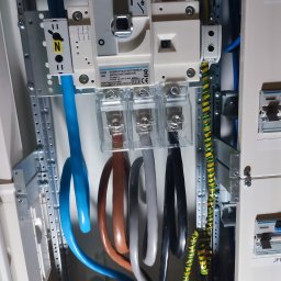 Damian Korzycki - Rozdzielnica elektryczna z bezpiecznikiem i podłączonymi przewodami o różnych kolorach, w tym przewód uziemiający w żółto-zielonej izolacji. Widoczne elementy montażowe.