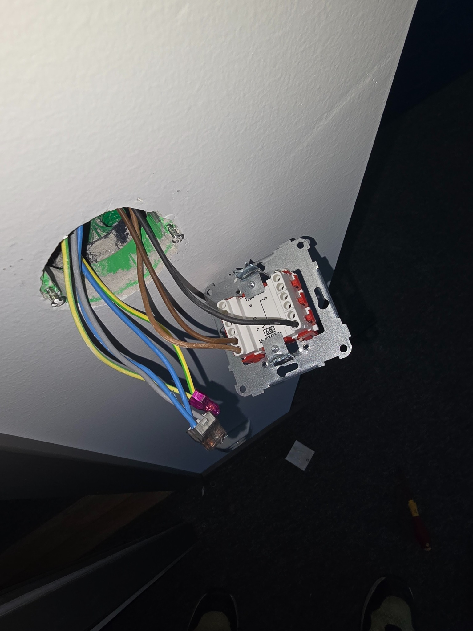 Widok z dołu na gniazdo elektryczne w trakcie montażu w ścianie, z widocznymi przewodami i elementami mocującymi. W tle fragment podłogi i narzędzie.