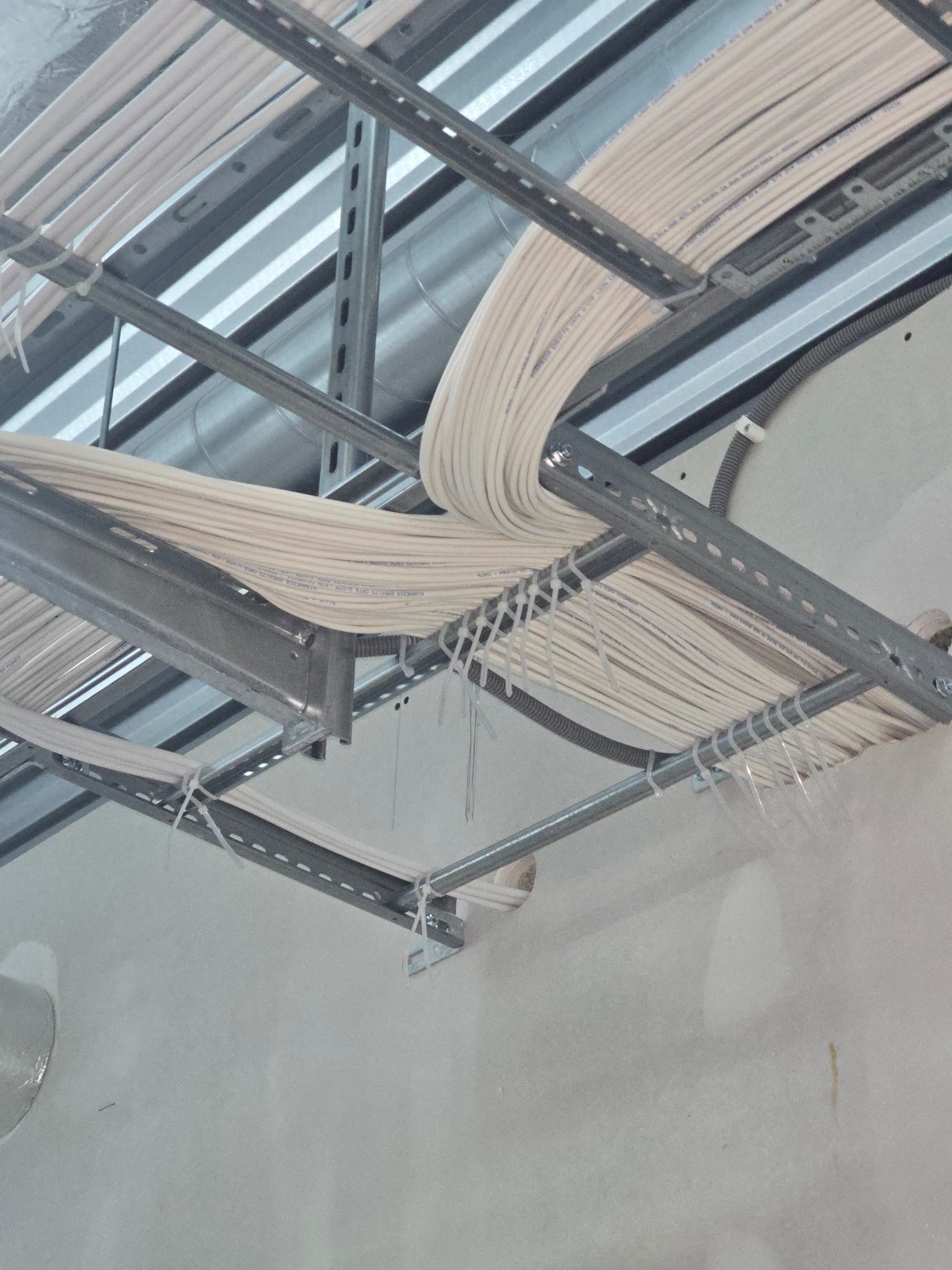 Białe kable elektryczne ułożone w metalowych korytkach pod sufitem, zabezpieczone plastikowymi opaskami. Widok z dołu na instalację w trakcie montażu.