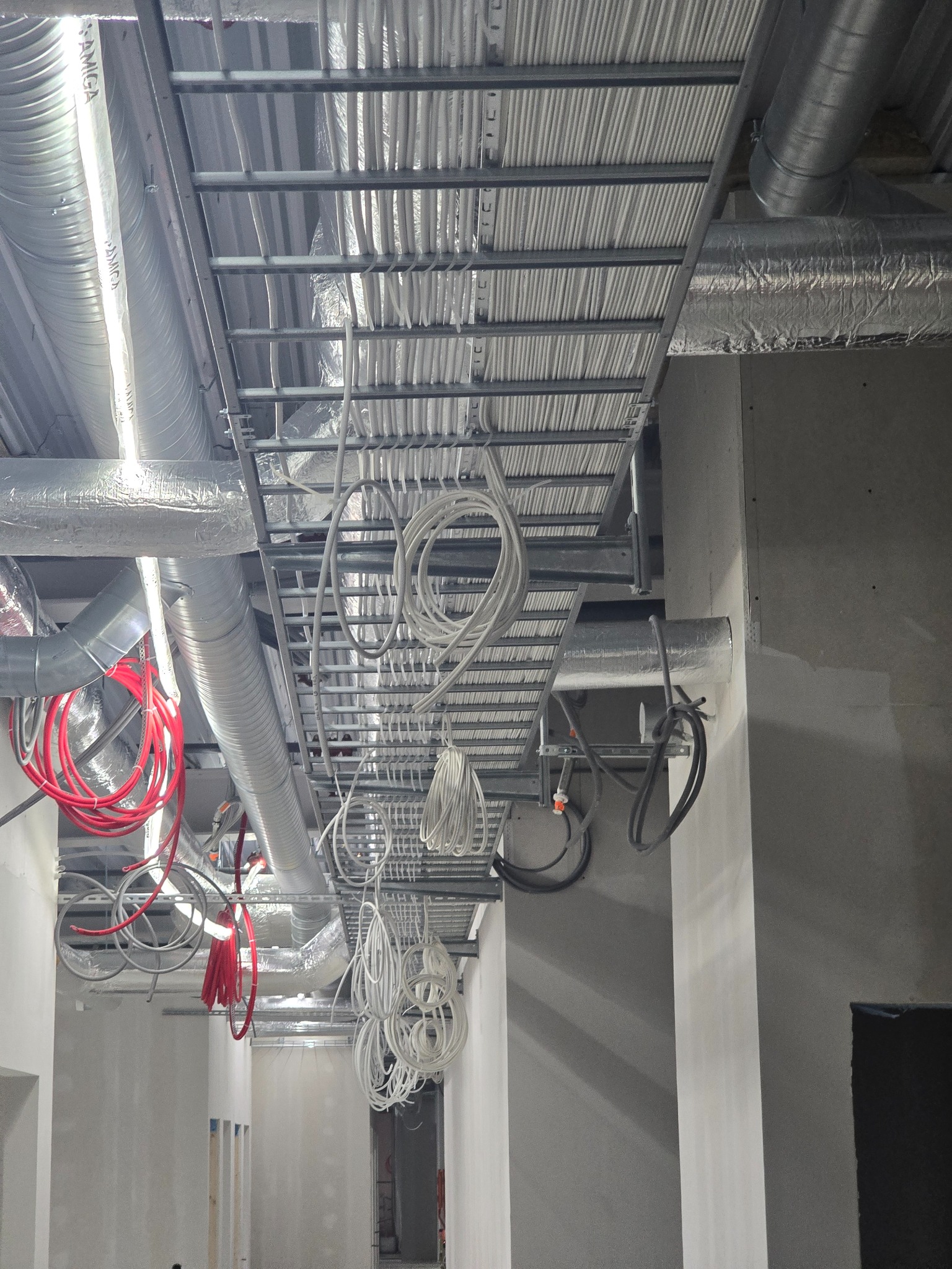 Metalowa konstrukcja pod sufitem z licznymi kablami i rurami wentylacyjnymi w budynku w trakcie wykończenia. Widoczne przewody ułożone w korytkach i zwisające pętle kabli.