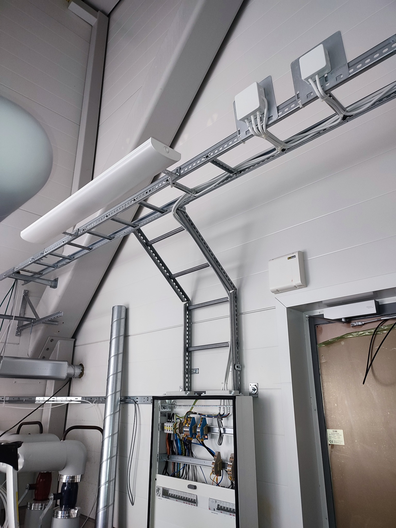 Szara instalacja elektryczna w hali z oświetleniem LED, korytkami kablowymi i rozdzielnicą. Widoczne przewody, zabezpieczenia i elementy montażowe.