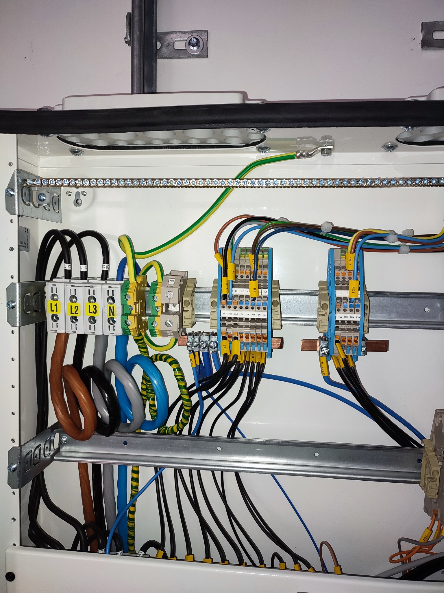 Wnętrze rozdzielnicy elektrycznej z widocznymi przewodami oznaczonymi kolorami, listwami zaciskowymi i szynami montażowymi. Przejrzysty układ kabli w białej obudowie.