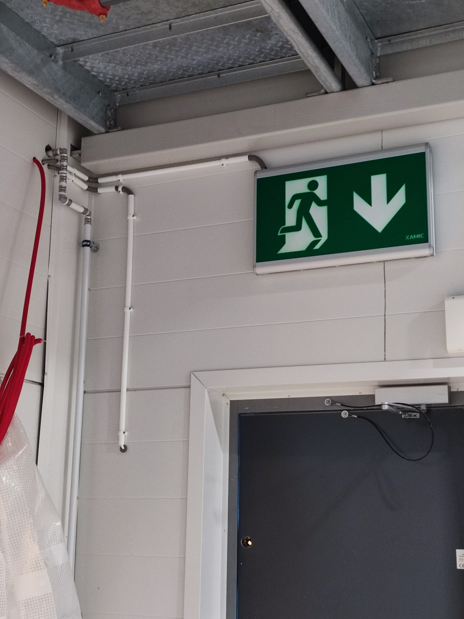 Zamontowany znak ewakuacyjny z zieloną ikoną i strzałką w dół, rury instalacji pod sufitem, szara ściana panelowa i górna część drzwi.