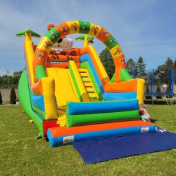 Dmuchaniec Nadarzyn - Kolorowa dmuchana zjeżdżalnia Tygrys na trawie, z małą dziewczynką w pobliżu. W tle zielone drzewa, trampolina i błękitne niebo.