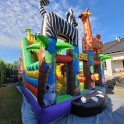 Kolorowy dmuchaniec Zoo z żyrafą i zebrą na tle błękitnego nieba, rozstawiony na trawniku. Widoczne detale: palmy, lew, słoń. Całość w jasnym świetle.