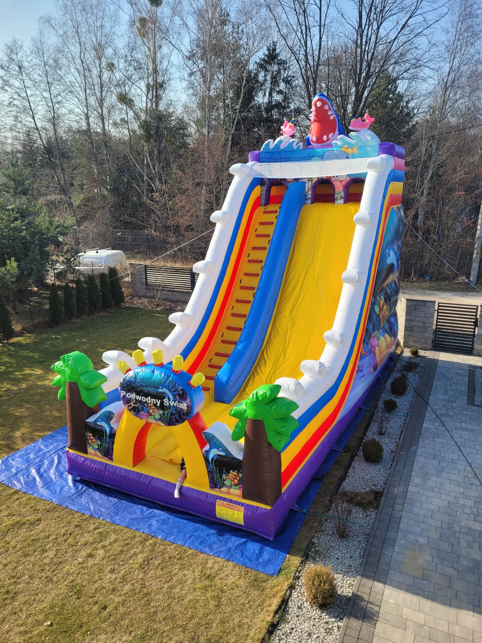 Kolorowa dmuchana zjeżdżalnia 'Podwodny Świat' na trawie, z motywami morskimi i rekinem na szczycie, ustawiona na niebieskiej plandece w ogrodzie.