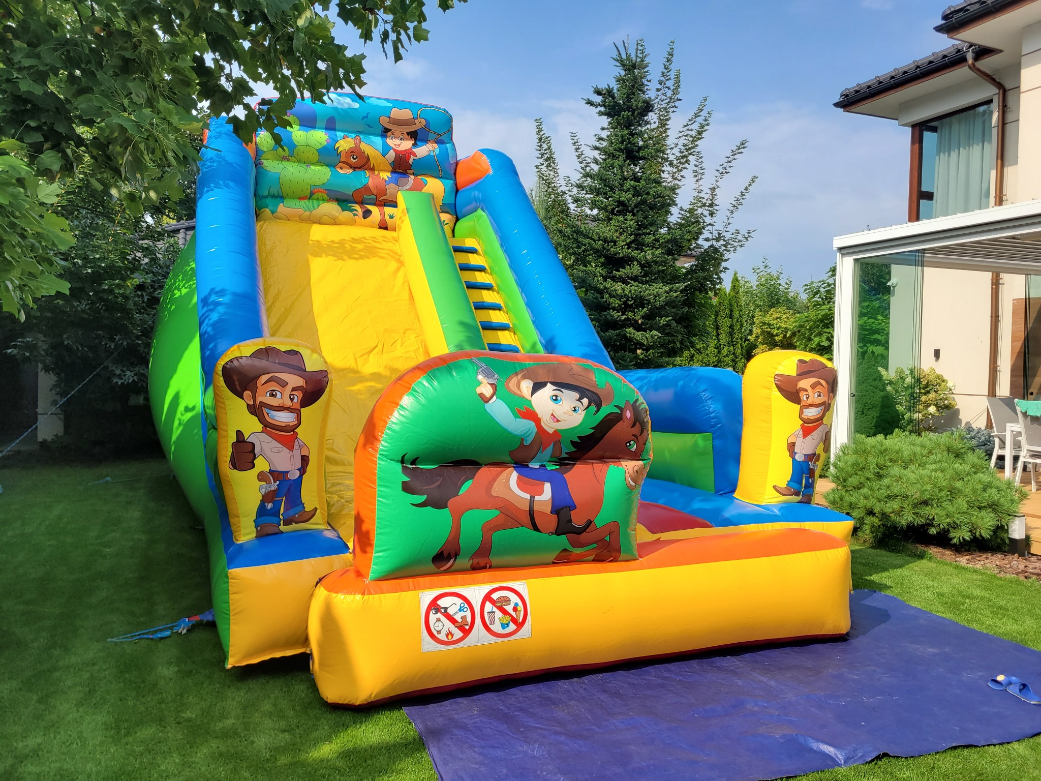 Kolorowa, dmuchana zjeżdżalnia w stylu western z grafiką kowbojów, ustawiona na trawniku przed domem. Widoczne drzewa i fragment budynku w tle.
