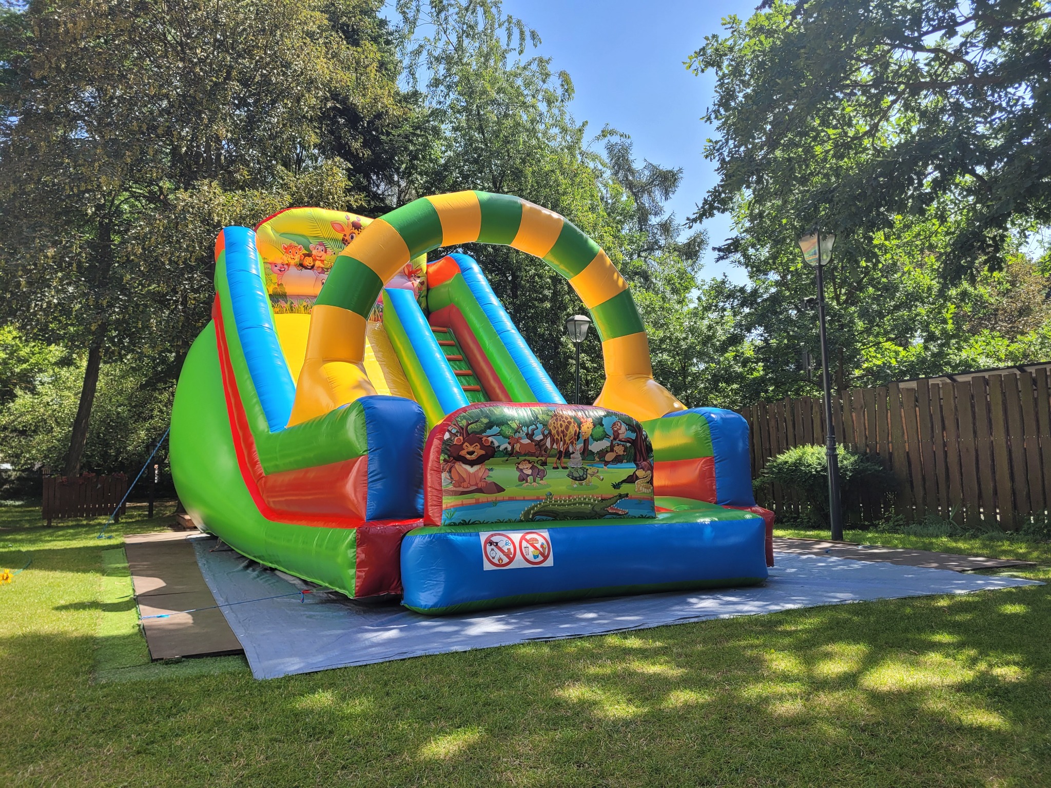Kolorowa, dmuchana zjeżdżalnia Safari z motywem dżungli, ustawiona na trawie w ogrodzie. Widoczne drzewa i drewniany płot w tle.