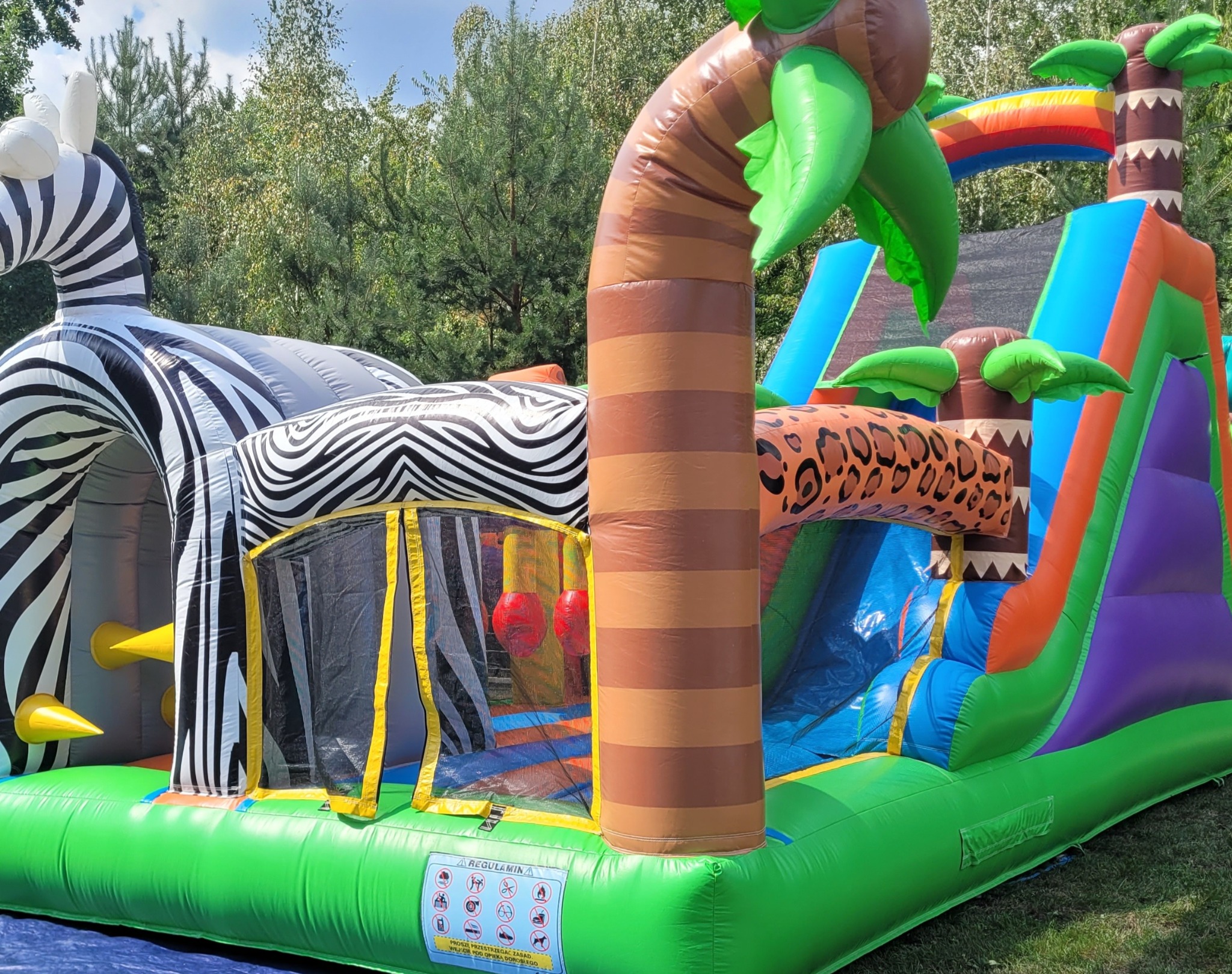 Dmuchany plac zabaw w stylu safari z motywem zebry, palmy i zjeżdżalnią. Kolorowa konstrukcja na trawie, idealna atrakcja dla dzieci na imprezy plenerowe.