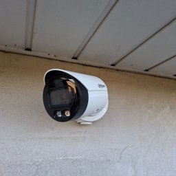 Smart Elektro Dawid Kowalski - Zamontowana kamera monitoringu Dahua na szarej ścianie pod okapem. Widoczne diody IR. Kąt ujęcia od dołu, podkreślający dyskretny montaż.