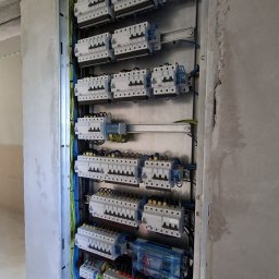 Smart Elektro Dawid Kowalski - Rozdzielnia elektryczna z licznymi bezpiecznikami i okablowaniem, zamontowana w surowej ścianie. Widoczne przewody w różnych kolorach i elementy zabezpieczające.