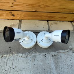 Smart Elektro Dawid Kowalski - Dwie białe kamery monitoringu Dahua zamontowane na szarej, chropowatej ścianie pod drewnianą okładziną. Kamery skierowane w różne strony, widoczne śruby montażowe.