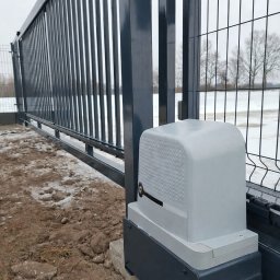 Smart Elektro Dawid Kowalski - Szara, przesuwna brama z automatyką na tle zaśnieżonego terenu. Widoczny mechanizm napędowy bramy, szyny i ogrodzenie z siatki.