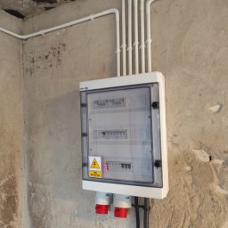 Smart Elektro Dawid Kowalski - Zamknięta skrzynka elektryczna z widocznymi bezpiecznikami, zamontowana na surowej ścianie. Nad nią białe rury instalacyjne. Dwa gniazda siłowe z czerwonymi wtykami na dole.