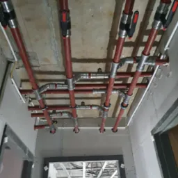 Czerwone rury instalacji hydraulicznej pod sufitem z płyt gipsowo-kartonowych. Widoczne metalowe łączenia i zawory. W tle konstrukcja sufitu podwieszanego.