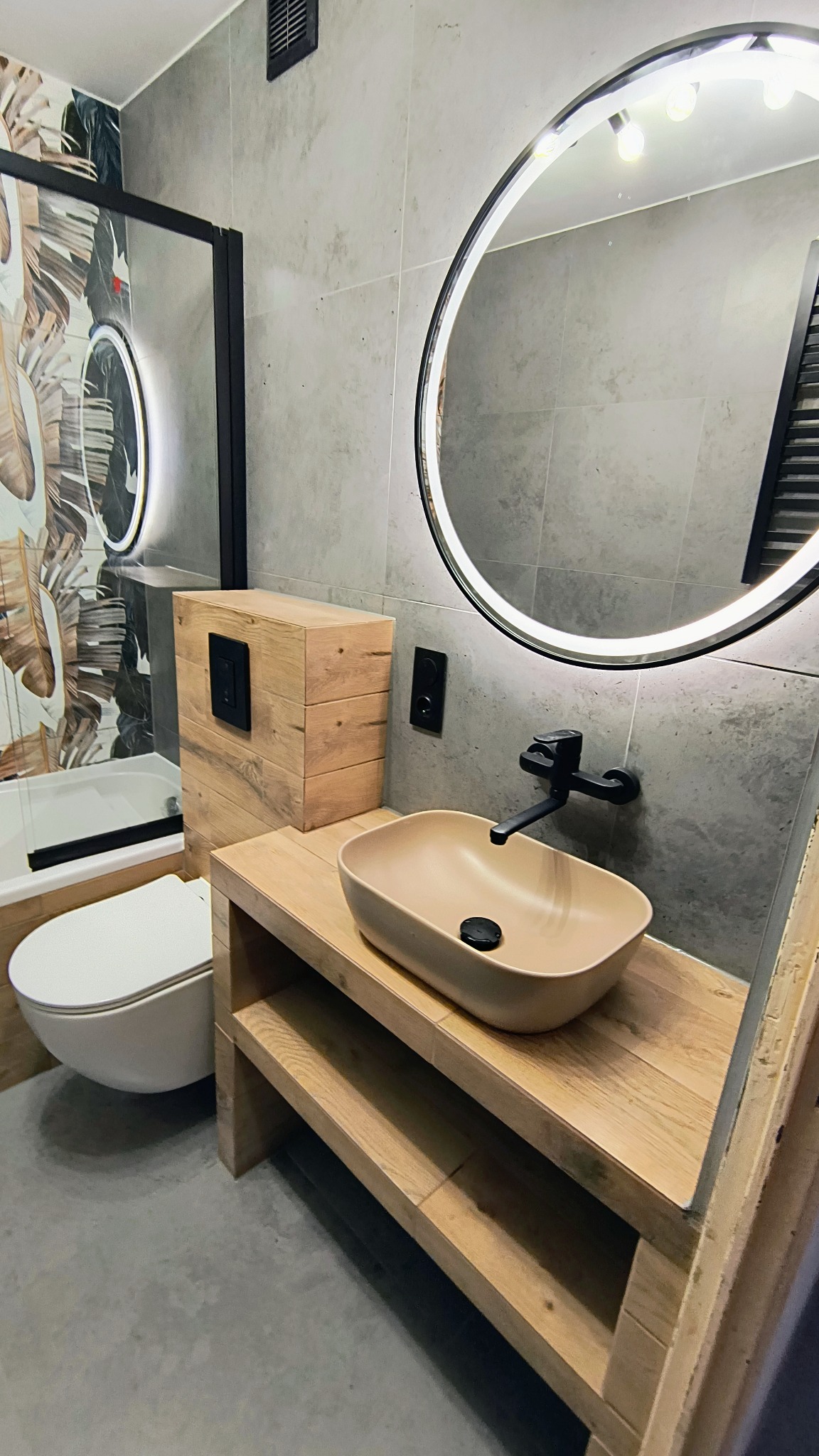 Nowoczesna łazienka z beżową umywalką na drewnianym blacie, okrągłe lustro z podświetleniem LED, szare płytki i toaleta w stylu skandynawskim.