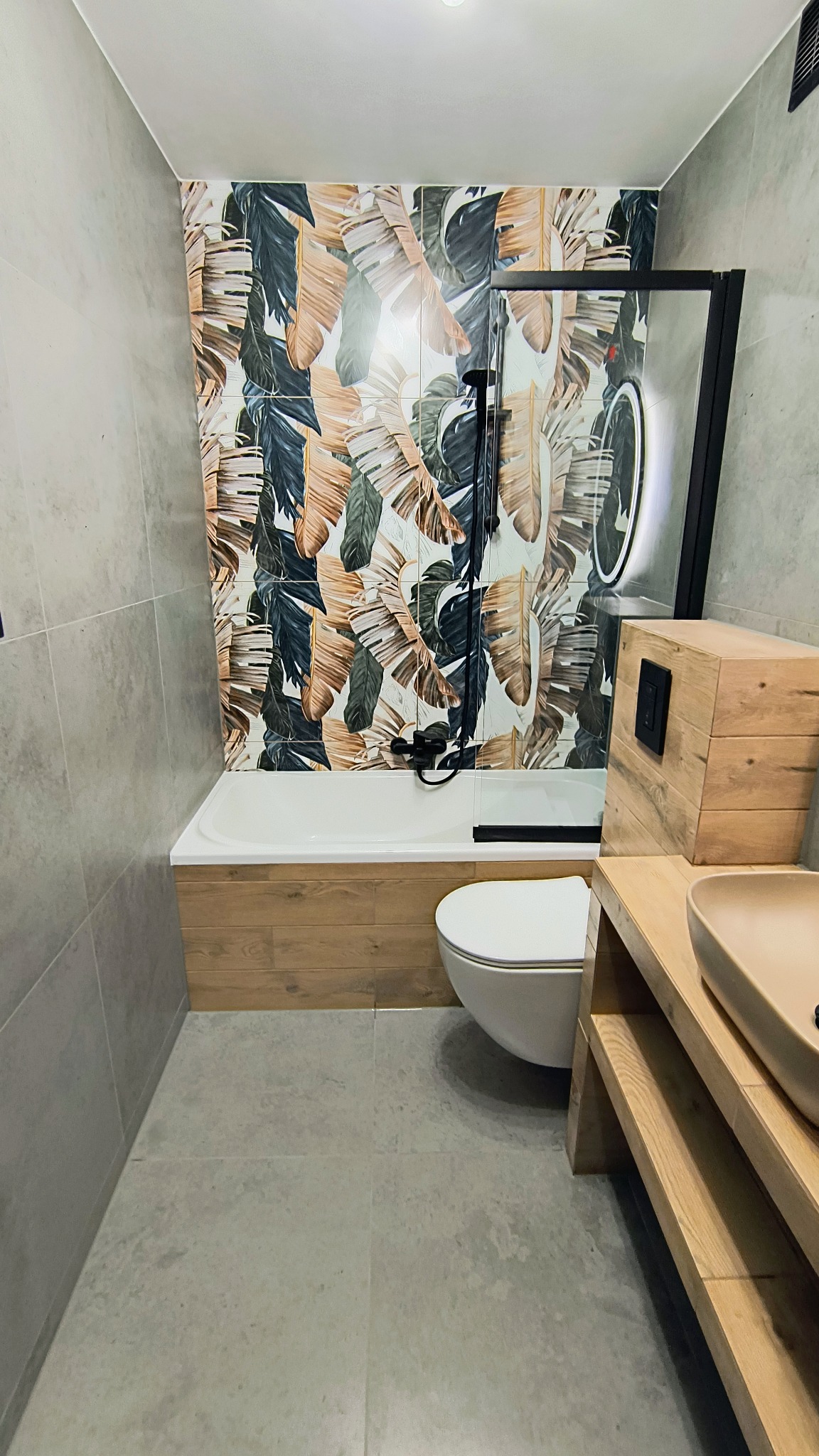 Nowoczesna łazienka z wanną obudowaną drewnem i ścianą w tropikalne liście. Umywalka na drewnianym stelażu, szara podłoga i ściany. Czarna armatura.