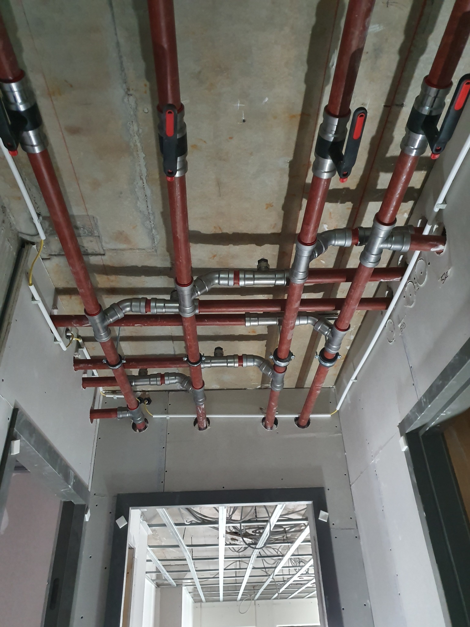 Czerwone rury instalacji hydraulicznej pod sufitem z płyt gipsowo-kartonowych. Widoczne metalowe łączenia i zawory. W tle konstrukcja sufitu podwieszanego.