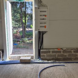 ELEKTROINSTALACJE ARTUR KOLSKI - Rozdzielnia elektryczna z przewodami w nowym budynku. Widoczne kable w korytach i peszlach, surowy beton. Okno z widokiem na zieleń w tle.