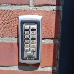 ELEKTROINSTALACJE ARTUR KOLSKI - Szary panel numeryczny domofonu Vidos zamontowany na czerwonej, ceglanej ścianie budynku, oświetlony naturalnym światłem słonecznym.