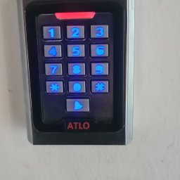 ELEKTROINSTALACJE ARTUR KOLSKI - Zamontowany, podświetlany panel domofonu ATLO z klawiaturą numeryczną na jasnej ścianie, widok z bliska.