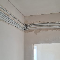 ELEKTROINSTALACJE ARTUR KOLSKI - Białe kable elektryczne ułożone równolegle pod sufitem w surowym wnętrzu. Widoczne mocowania kabli do ściany. Ściany w trakcie prac wykończeniowych.