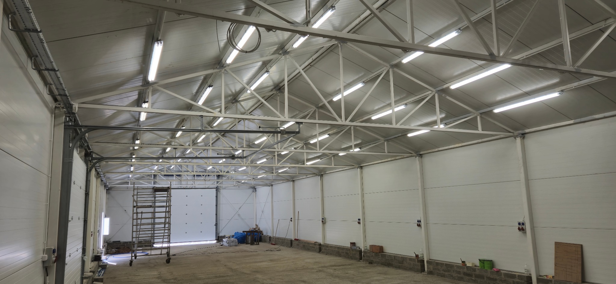 Wnętrze hali z oświetleniem LED zamontowanym pod metalową konstrukcją dachu. Widoczne kable i przewody w korytkach. Ściany wyłożone panelami.