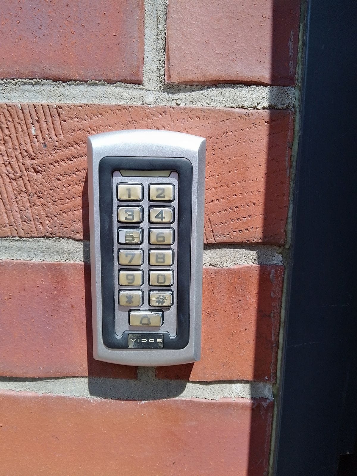 Szary panel numeryczny domofonu Vidos zamontowany na czerwonej, ceglanej ścianie budynku, oświetlony naturalnym światłem słonecznym.